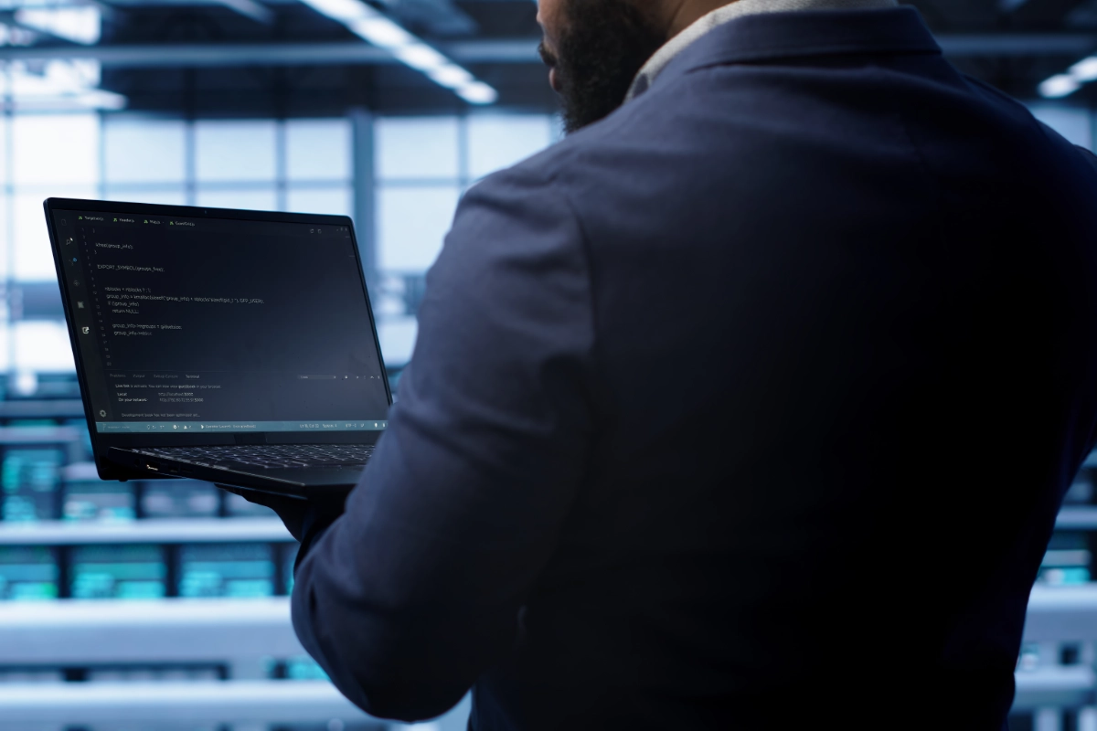managed-it-img-1 Person in a dark suit holding a laptop displaying code in a blue-toned office setting.