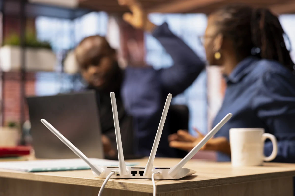 network-design-voip-img-1 Wi‑Fi router with four white antennas on a desk, office backdrop with two people arguing in the background.