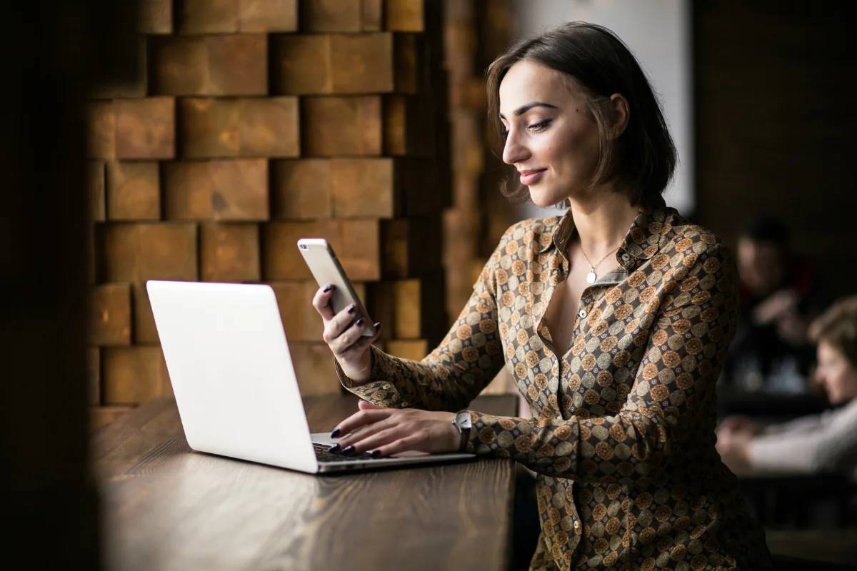 network-design-voip-img-4 Woman in a patterned shirt at a wooden counter, typing on a laptop and checking her phone in a cafe.